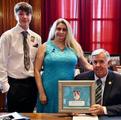 GOVERNOR PARSON SIGNED ‘RANDOM ACTS OF KINDNESS’ BILL INTO LAW – Governor Mike Parson signed Senate Bill 72 on Tuesday, July 13th, 2021 in Jefferson City, Mo. Joining the Governor for the signing are Brady Thompson, left and Lela Thompson as he holds a photo of Shayley Akers. The “Random Acts of Kindness Day” has been observed locally in Washington County for the past few years and is now officially recognized by the State of Missouri, to be honored on the last day of August, prior to September’s Suicide Awareness Month, annually. Lela thanked the Governor for the important recognition. (State Photo)
